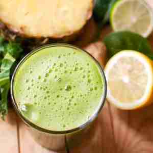 PCOS Breakfast idea-Glass with frothy green smoothie in the forefront with a background of pineapple, lemons and greens.