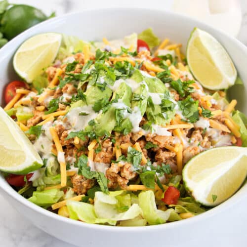 white bowl filled with a PCOS-friendly taco salad topped with lime wedges, shredded cheese and cilantro yogurt dressing.